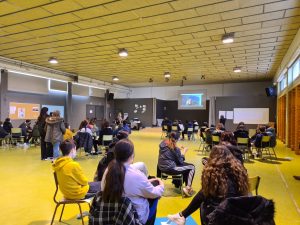 Teenagers in Spain sitting in a big gymnasium hall. They are listening to a speaker.