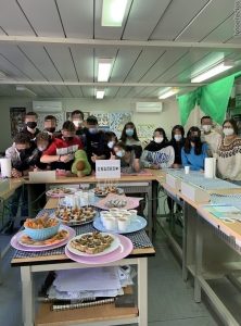 A large group of teenage scientists standing around tables with the snacks they prepared. All of their faces are blurred out for safety.