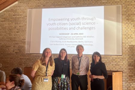 Four people stand in front of a screen that says 'Empowering Youth through youth citizen (social) science: possibilities and challenges. Cathrine is on the left wearing a yellow dress and has blond hair. Claire is standing beside her. She has brown hair and is wearing a black top and a purple/green/yellow vibrant patterned skirt. Michael is beside Claire and he has short grey hair and is wearing a light grey/pink shirt with black trousers. Eglė is beside Michael, standing on the far right, wearing a black dress with dark brown hair. Everyone is smiling happily