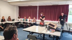 Claire Murray is standing in a conference room with lots of people seated listening to her talk. She is a white woman with brunette hair wearing a black top and a pink skirt