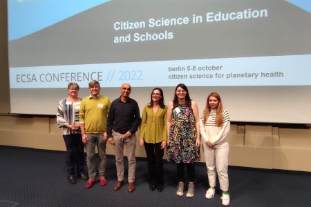 Six people standing in a line in front of a large display screen that says 'Citizen Science in Education and Schools. There are four women and two men, and all are standing and smiling